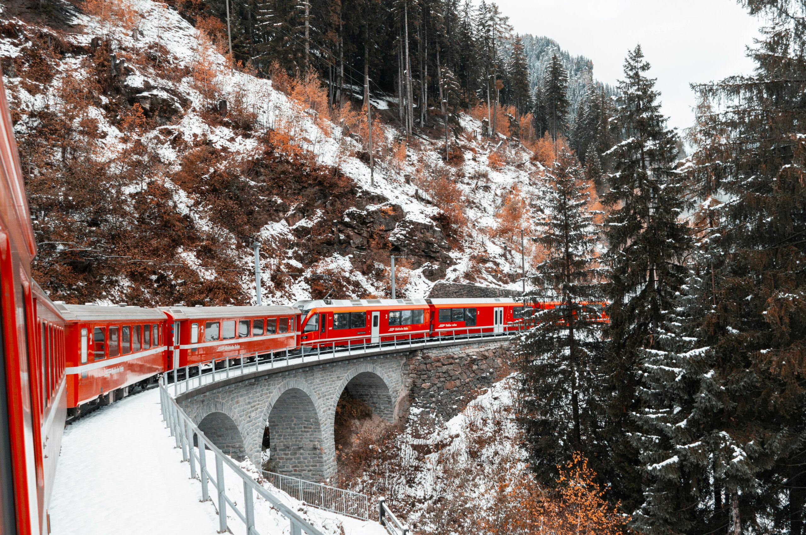 A vibrant red train crosses a snowy coniferous landscape on a scenic mountain bridge in winter.