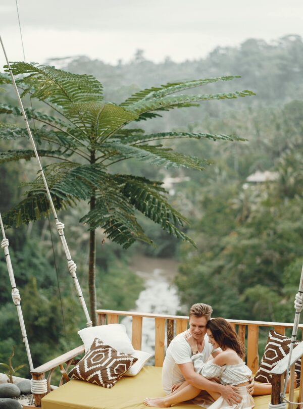 Side view full length barefoot stylish couple bonding and relaxing on cozy hanging swing together during romantic holiday in tropical countryside