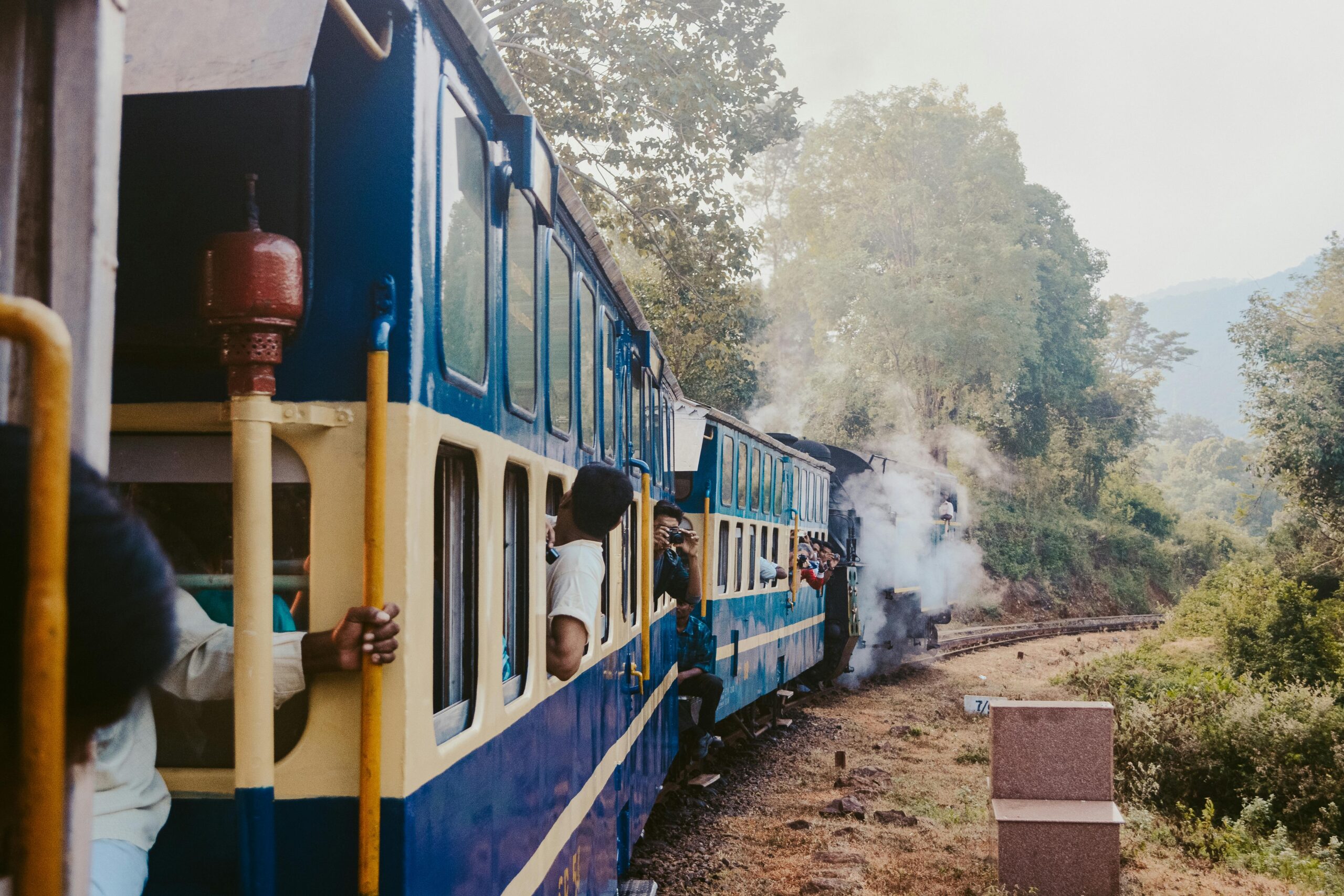 A vintage train with passengers travels through a scenic, forested area, emitting steam.